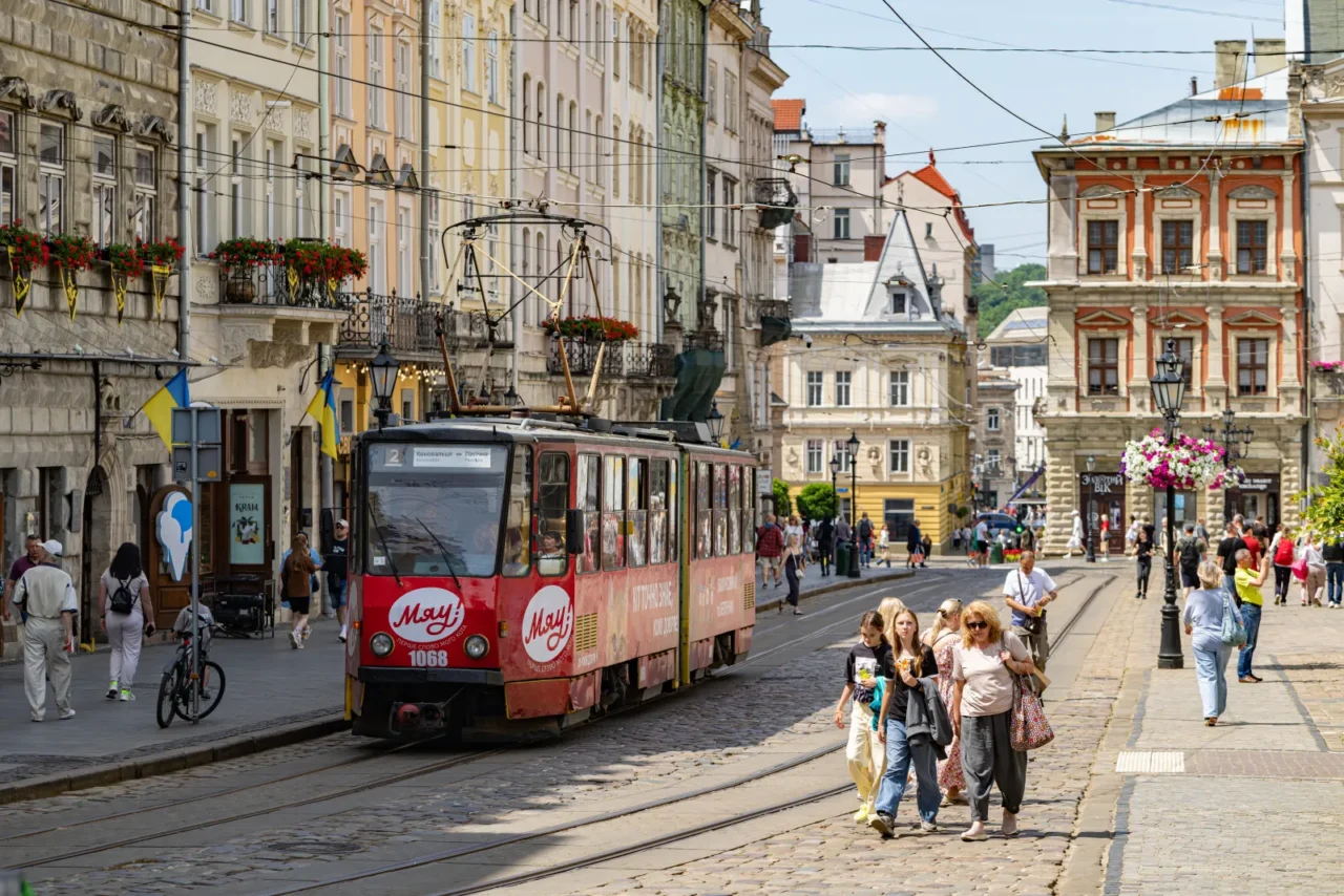 Historic tram and pedestrians on a cobbled street in Lviv, Ukraine, with colorful old buildings and Ukrainian flags.